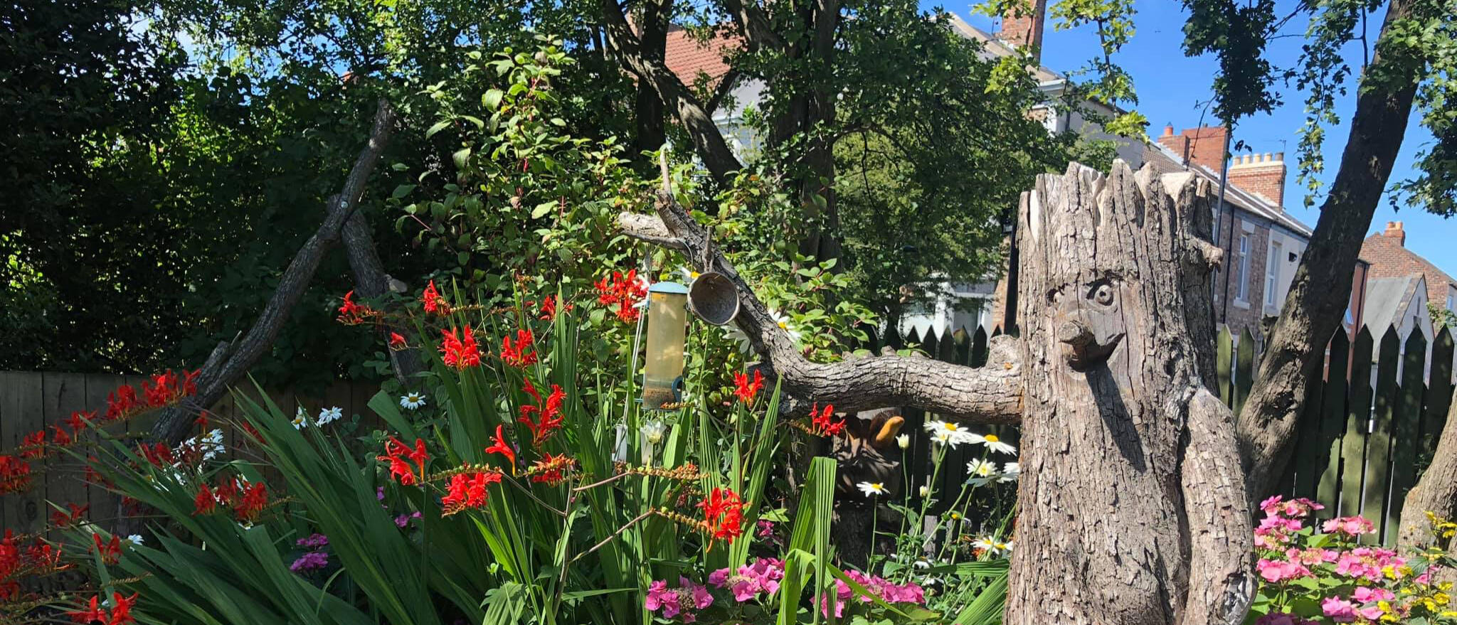 A tree sculpture in the garden, carved with a kind face and hair. A bird feeder hangs from its branch above a bed of plants.